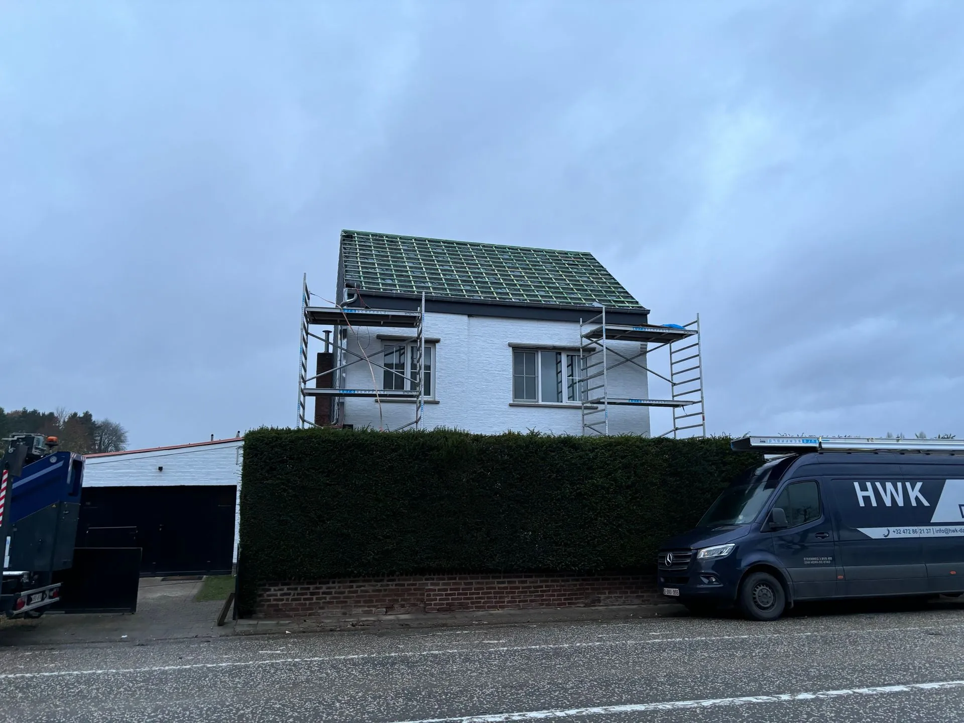 Voorgevel van een woning tijdens dak- en gevelwerken, met groene onderdakfolie en steigers.