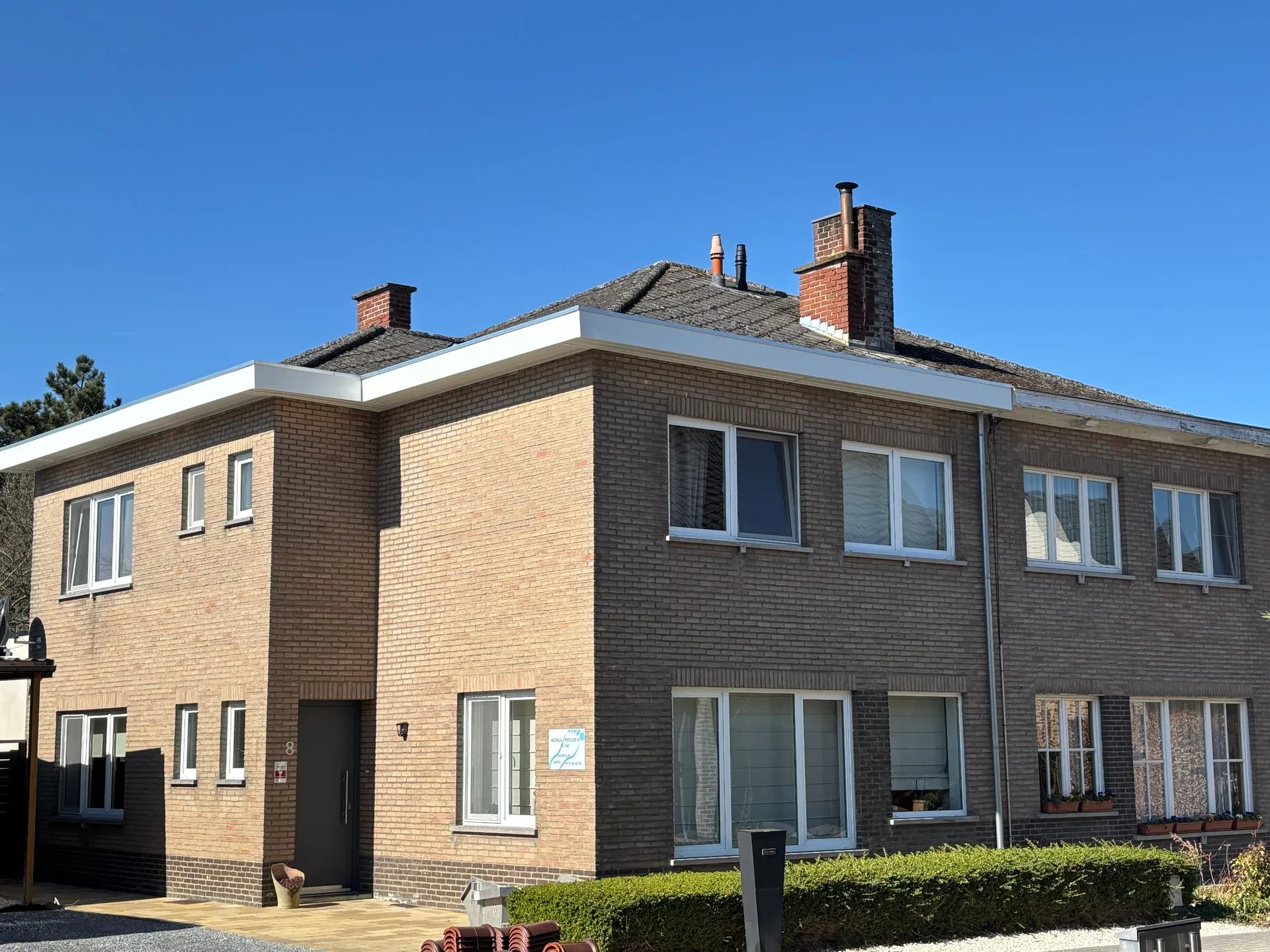 Straatzijde van een hoekwoning in Hoegaarden met een antracietkleurig pannendak en twee schoorstenen.